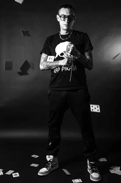 Inoki в чёрной футболке RAP PIRATA среди летящих карт, чёрно-белое фото